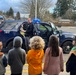 A police officer is conducting an educational demonstration for a group of children in a parking lot, showing them a police patrol SUV. The officer is standing beside the open door of a dark navy blue police vehicle with its emergency lights activated. This appears to be a community outreach or school visit event where children are learning about law enforcement.
