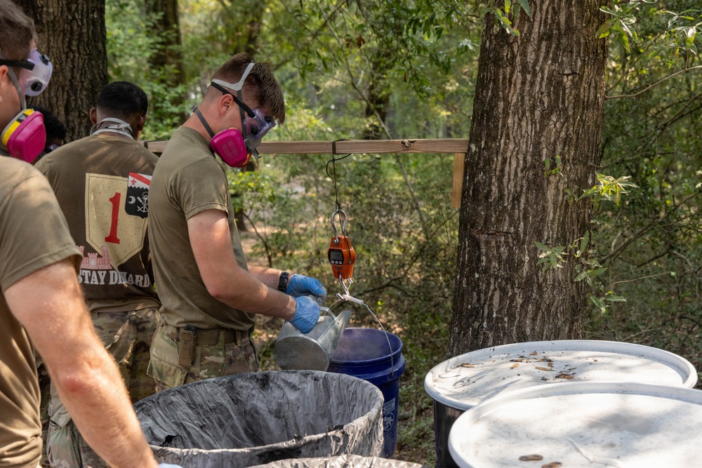 Military personnel in olive drab t-shirts and camouflage pants are conducting what appears to be a hazardous materials handling exercise in a wooded outdoor setting. The soldiers are wearing respiratory protection (half-face respirators) and safety goggles while working with containers and a hanging scale. The shirt visible on the left reads 'STAY DIEHARD' with what appears to be a unit patch featuring the number '1'.