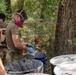 Military personnel in olive drab t-shirts and camouflage pants are conducting what appears to be a hazardous materials handling exercise in a wooded outdoor setting. The soldiers are wearing respiratory protection (half-face respirators) and safety goggles while working with containers and a hanging scale. The shirt visible on the left reads 'STAY DIEHARD' with what appears to be a unit patch featuring the number '1'.