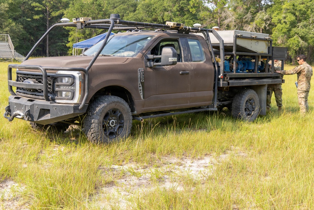 A military-configured Ford F-250/F-350 Super Duty truck painted in matte brown/earth tone is parked in an open grassy field. The truck features a custom flatbed with mounted equipment, a roof rack with sensors or antennas, and a heavy-duty front bumper with a winch. A soldier in camouflage uniform is standing at the rear of the vehicle, appearing to inspect or operate the bed-mounted equipment.