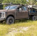 A military-configured Ford F-250/F-350 Super Duty truck painted in matte brown/earth tone is parked in an open grassy field. The truck features a custom flatbed with mounted equipment, a roof rack with sensors or antennas, and a heavy-duty front bumper with a winch. A soldier in camouflage uniform is standing at the rear of the vehicle, appearing to inspect or operate the bed-mounted equipment.