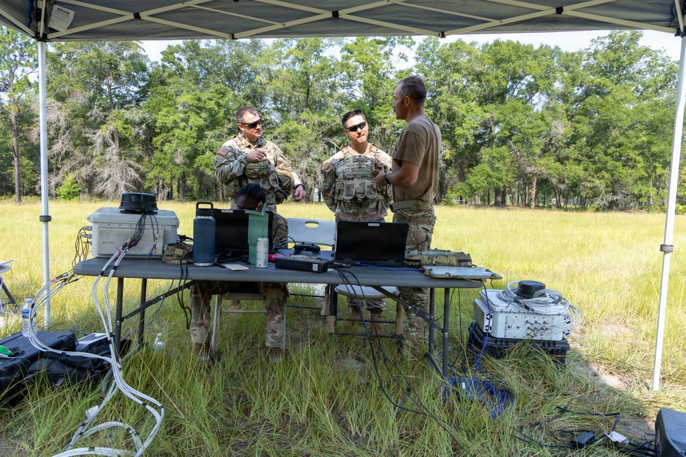 Three military personnel are gathered under a portable canopy tent in an open field, standing behind a table equipped with laptops, communication equipment, and various electronic devices. Two individuals are wearing full camouflage uniforms with tactical vests, while the third wears a plain tan/brown t-shirt. The setup appears to be a field command or communications station during a training exercise or field operation.