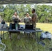 Three military personnel are gathered under a portable canopy tent in an open field, standing behind a table equipped with laptops, communication equipment, and various electronic devices. Two individuals are wearing full camouflage uniforms with tactical vests, while the third wears a plain tan/brown t-shirt. The setup appears to be a field command or communications station during a training exercise or field operation.