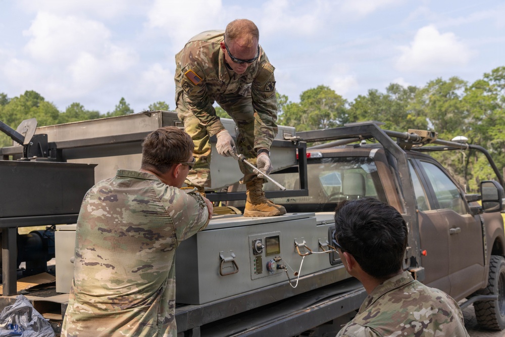 Three military personnel in US Army OCP (Operational Camouflage Pattern) uniforms are working on equipment mounted in the bed of a tactical utility truck. One soldier is standing on the truck bed using a tool on a gray equipment case, while two others observe and assist from ground level. The setting appears to be an outdoor training or operational area with trees visible in the background.