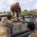 Three military personnel in US Army OCP (Operational Camouflage Pattern) uniforms are working on equipment mounted in the bed of a tactical utility truck. One soldier is standing on the truck bed using a tool on a gray equipment case, while two others observe and assist from ground level. The setting appears to be an outdoor training or operational area with trees visible in the background.