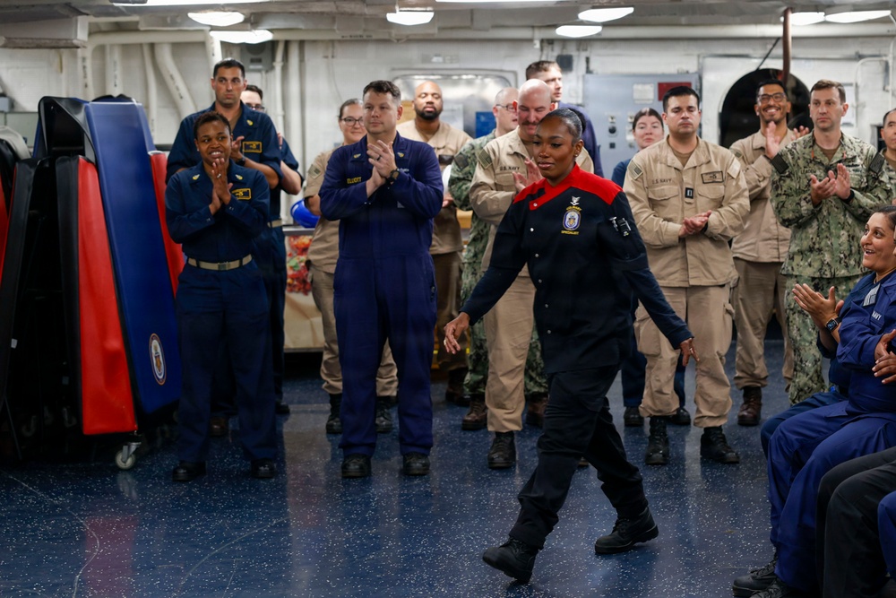 A military ceremony or recognition event is taking place aboard what appears to be a U.S. Navy vessel. A female sailor wearing a distinctive dark uniform with red accents walks forward while surrounded by applauding fellow service members in various Navy uniforms. The atmosphere appears celebratory, suggesting an award, promotion, or special recognition ceremony.