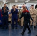 A military ceremony or recognition event is taking place aboard what appears to be a U.S. Navy vessel. A female sailor wearing a distinctive dark uniform with red accents walks forward while surrounded by applauding fellow service members in various Navy uniforms. The atmosphere appears celebratory, suggesting an award, promotion, or special recognition ceremony.