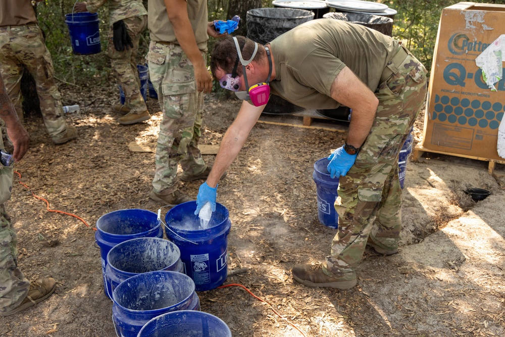 Military personnel in multicam/OCP uniforms are conducting what appears to be a field training or operational exercise involving blue buckets from Lowe's. The central figure is wearing a half-face respirator mask and blue nitrile gloves while handling materials in the buckets, suggesting work with potentially hazardous substances. The setting is an outdoor wooded area with additional equipment and containers visible in the background.