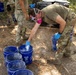 Military personnel in multicam/OCP uniforms are conducting what appears to be a field training or operational exercise involving blue buckets from Lowe's. The central figure is wearing a half-face respirator mask and blue nitrile gloves while handling materials in the buckets, suggesting work with potentially hazardous substances. The setting is an outdoor wooded area with additional equipment and containers visible in the background.