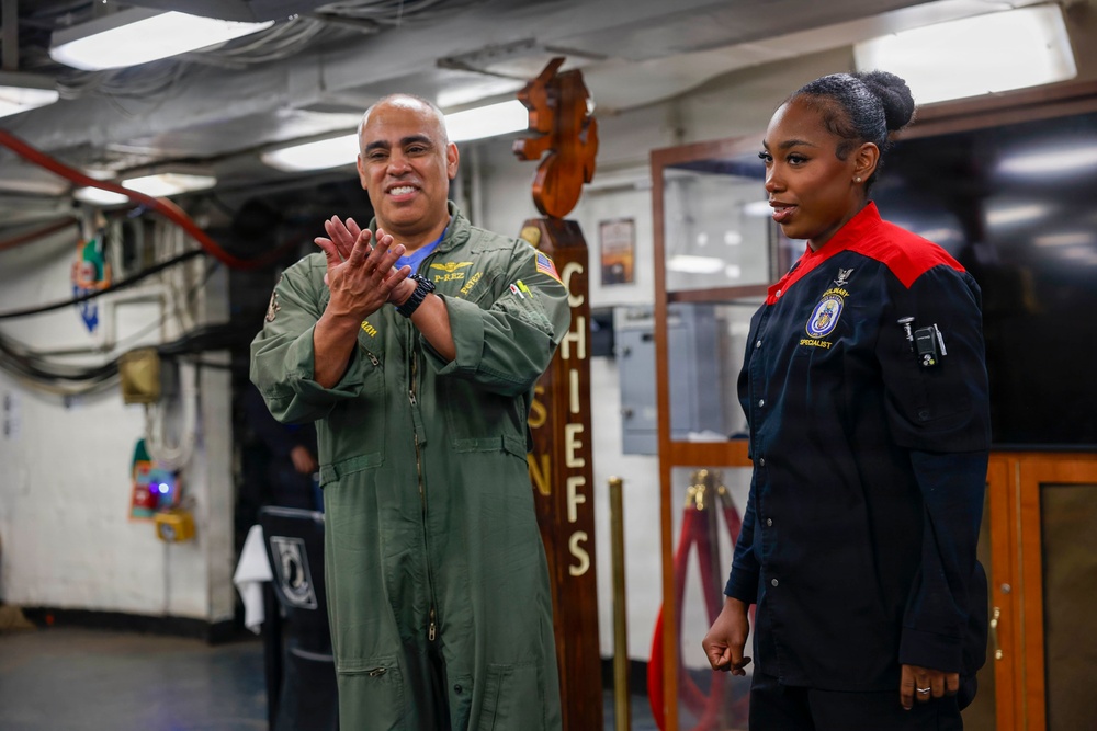 Two military personnel are standing inside what appears to be a naval vessel's Chiefs mess area. A male service member in an olive green flight suit is clapping, while a female service member in a dark navy uniform with red accents stands beside him. The setting suggests a formal or ceremonial occasion within a U.S. Navy environment.