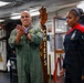 Two military personnel are standing inside what appears to be a naval vessel's Chiefs mess area. A male service member in an olive green flight suit is clapping, while a female service member in a dark navy uniform with red accents stands beside him. The setting suggests a formal or ceremonial occasion within a U.S. Navy environment.