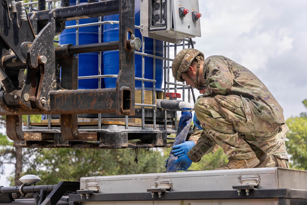 A soldier in camouflage uniform and blue protective gloves is working with a liquid transfer system, appearing to connect or manage a hose/pipe fitting on what looks like a water or chemical distribution setup. The equipment includes large blue IBC (Intermediate Bulk Container) tanks mounted on a pallet frame with a metal cage, along with an electrical control panel. The setting appears to be an outdoor military or disaster relief operation.