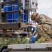 A soldier in camouflage uniform and blue protective gloves is working with a liquid transfer system, appearing to connect or manage a hose/pipe fitting on what looks like a water or chemical distribution setup. The equipment includes large blue IBC (Intermediate Bulk Container) tanks mounted on a pallet frame with a metal cage, along with an electrical control panel. The setting appears to be an outdoor military or disaster relief operation.