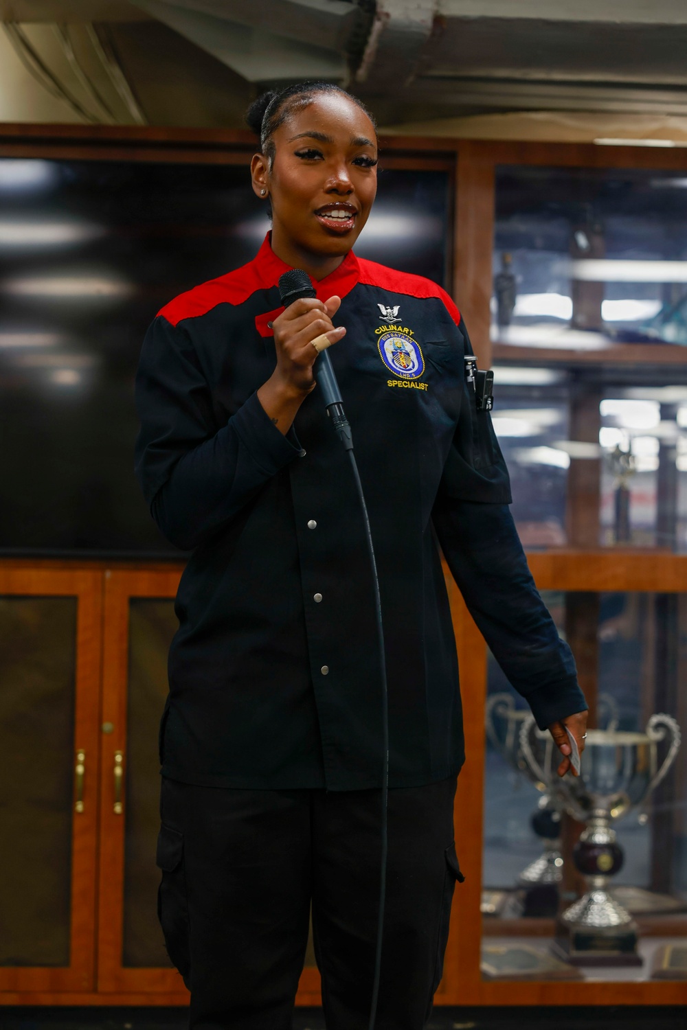 A woman in a U.S. Navy Culinary Specialist uniform is speaking into a microphone, likely at a formal or ceremonial event. Her uniform is black with red accents and bears an embroidered patch reading 'Culinary Specialist' along with a ship's crest. Trophy cups are visible in the background, suggesting an awards or recognition setting.