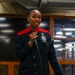 A woman in a U.S. Navy Culinary Specialist uniform is speaking into a microphone, likely at a formal or ceremonial event. Her uniform is black with red accents and bears an embroidered patch reading 'Culinary Specialist' along with a ship's crest. Trophy cups are visible in the background, suggesting an awards or recognition setting.