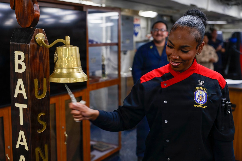 A U.S. Navy Culinary Specialist is ringing a brass ship's bell mounted on a wooden post bearing the letters 'BATAN' (likely USS Bataan). She is wearing a distinctive black and red culinary uniform with a ship's crest patch identifying her role. Several other personnel are visible in the background in a shipboard interior setting.