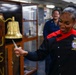 A U.S. Navy Culinary Specialist is ringing a brass ship's bell mounted on a wooden post bearing the letters 'BATAN' (likely USS Bataan). She is wearing a distinctive black and red culinary uniform with a ship's crest patch identifying her role. Several other personnel are visible in the background in a shipboard interior setting.
