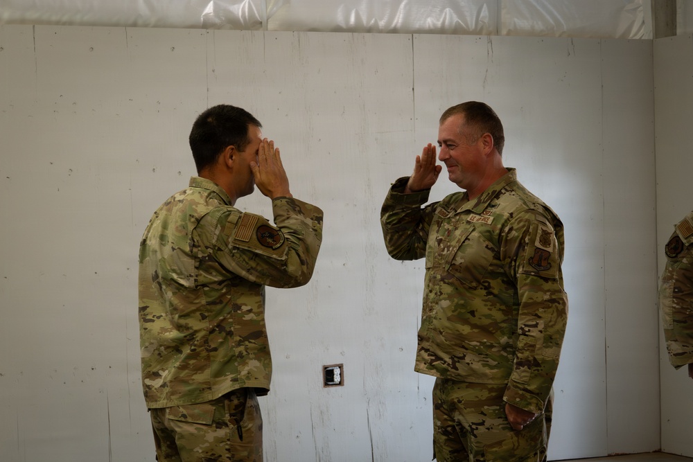 Two military personnel in OCP (Operational Camouflage Pattern) uniforms are exchanging salutes facing each other indoors. Both individuals wear U.S. Air Force uniforms with patches visible on their sleeves. The setting appears to be a temporary or field facility with white paneled walls.