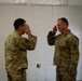 Two military personnel in OCP (Operational Camouflage Pattern) uniforms are exchanging salutes facing each other indoors. Both individuals wear U.S. Air Force uniforms with patches visible on their sleeves. The setting appears to be a temporary or field facility with white paneled walls.