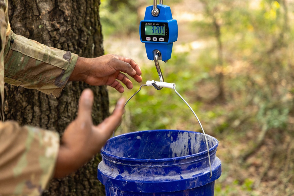 A person wearing military camouflage attire is using a blue digital crane scale to weigh a blue plastic bucket outdoors near a tree. The crane scale displays a reading of 32.05 on its green LCD screen. The setting appears to be a wooded or forested area.