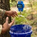 A person wearing military camouflage attire is using a blue digital crane scale to weigh a blue plastic bucket outdoors near a tree. The crane scale displays a reading of 32.05 on its green LCD screen. The setting appears to be a wooded or forested area.