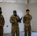 Three military personnel in camouflage uniforms are gathered indoors in what appears to be a formal ceremony or briefing. The central figure is reading from a dark folder, while the other two stand at attention on either side. The personnel appear to represent different military branches or nations based on their distinct uniform patterns and headgear.