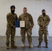 Three military personnel are posed together in what appears to be a formal ceremony or award presentation inside a building. The central figure is holding an open blue folder containing an official document, suggesting a commendation or certificate presentation. Two individuals wear Canadian Armed Forces uniforms with Canadian flag patches, while the center individual wears a U.S. military multicam uniform.