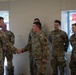 A group of military personnel in OCP (Operational Camouflage Pattern) uniforms are gathered inside what appears to be a building under construction or renovation. One individual on the left is shaking hands with another service member, suggesting a formal greeting, recognition, or introduction ceremony. The setting appears to be an informal indoor military event.