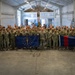 A large group of military personnel in camouflage uniforms pose for a formal group photograph inside what appears to be a military hangar or maintenance facility. Two flags are prominently displayed: the Indiana State flag (blue with torch and stars) on the left, and what appears to be a Canadian military unit flag (dark blue with red stripe) on the right. The group includes both American and Canadian military members, suggesting a joint training exercise or bilateral military event.
