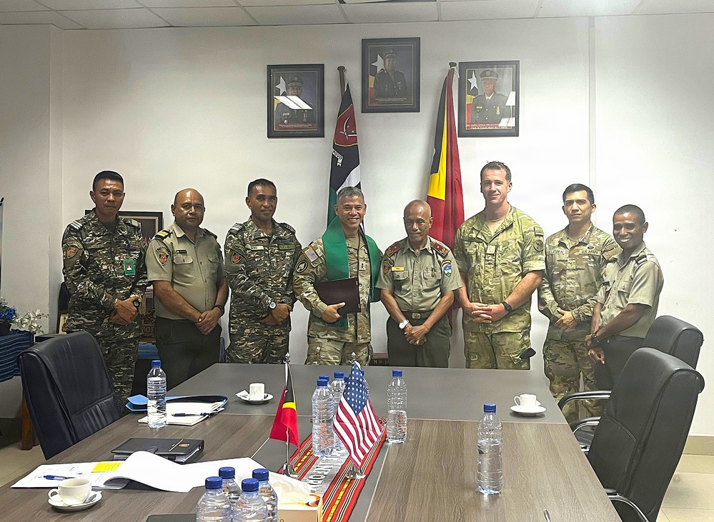 A formal military meeting or signing ceremony between personnel from what appears to be Timor-Leste armed forces and U.S. military representatives, taking place in an official conference room. Eight individuals in military uniforms are posed together behind a conference table, with one person holding what appears to be a document or folder. The setting includes national flags of Timor-Leste and the United States, along with framed portraits on the wall.