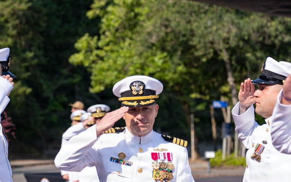 A U.S. Navy officer, likely of senior rank (Captain or Admiral based on insignia and gold oak leaf on cap), walks through a sideboy honors ceremony while being saluted by a row of naval personnel in white dress uniforms. The central figure displays numerous medals and ribbons on his white service dress uniform, suggesting a formal military ceremony such as a change of command, retirement, or arrival/departure honors. Wooden bollards line the ceremonial passageway, and additional uniformed personnel are visible in the background.