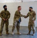 Three military personnel are gathered in what appears to be a temporary facility or hangar. A center figure in US Army MultiCam uniform is handing a document or folder to a Canadian Armed Forces member wearing a blue beret, while a second Canadian soldier wearing a dark blue Sikh dastar (turban) stands at ease to the left. The scene appears to depict a formal exchange or presentation ceremony.