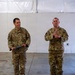 Two U.S. Air Force personnel in OCP (Operational Camouflage Pattern) uniforms stand in what appears to be a hangar or large indoor facility. The individual on the left, identified by name tape as 'CARPENTER,' is holding a blue folder and wearing a medal on his chest, suggesting a military awards ceremony. The individual on the right, identified as 'DOBSON,' is applauding.
