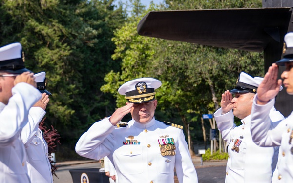 A U.S. Navy officer in white dress uniform walks a red carpet while being saluted by subordinate sailors on both sides. The central figure appears to be a senior officer, identifiable by the gold-braided cap and multiple service ribbons and medals on his uniform. This appears to be a formal military ceremony, likely a change of command, retirement, or official arrival/departure event.