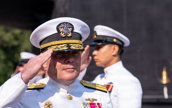 A formal U.S. Navy military ceremony is taking place outdoors, with multiple uniformed personnel rendering salutes. The foreground officer wears a white dress uniform adorned with numerous medals, ribbons, and gold insignia, indicating high senior rank. Additional naval personnel in various white dress uniforms are visible saluting in the background.