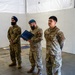 Three military personnel are standing inside a tent or temporary structure during what appears to be a formal ceremony or presentation. Two individuals are wearing Canadian Armed Forces uniforms with visible Canadian flag patches, while the third is wearing a U.S. Air Force uniform. The central figure is holding a blue certificate or award folder, suggesting a commendation or recognition event.