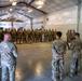 A large group of military personnel in camouflage uniforms are assembled inside a large hangar or warehouse-style facility. A smaller group of individuals, including what appears to be a Canadian Armed Forces member wearing a blue beret and a dark beard, are addressing the assembled troops. The setting suggests a formal briefing, ceremony, or unit formation.