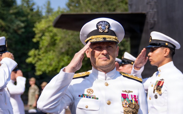 A formal U.S. Navy military ceremony is taking place outdoors, with multiple naval officers in white dress uniforms rendering salutes. The central figure, a senior officer distinguished by gold shoulder boards with multiple stars and an extensive array of medals and decorations, appears to be the focal point of the ceremony. A red carpet and ceremonial stanchions are visible, suggesting a change of command or high-level official event.