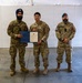 Three military personnel are posed together in what appears to be a formal recognition ceremony inside a tent or temporary structure. The center individual, wearing U.S. Air Force uniform, is holding a blue folder containing what appears to be a certificate or commendation letter. The two flanking individuals are wearing Canadian Armed Forces uniforms, identifiable by Canadian flag patches on their shoulders.