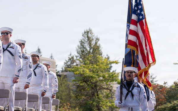 A U.S. Navy color guard ceremony is taking place outdoors, with sailors in dress white uniforms carrying the American flag and what appears to be a service flag. A formation of Navy personnel stands at attention alongside folding chairs, suggesting a formal military ceremony or commemoration event. The setting appears to be a military installation or memorial site, likely during daytime in a temperate season given the green foliage.