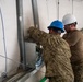 Two workers in military-style camouflage uniforms are performing electrical or mechanical installation work on a wall-mounted metal enclosure or junction box. One worker wears a blue hard hat and the other wears a white hard hat with safety glasses. They appear to be working inside a temporary structure with fabric/tent-style walls.