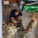 Three individuals, two in U.S. military OCP (Operational Camouflage Pattern) uniforms and one in civilian/mixed attire, are working together on a mechanical or HVAC unit on a concrete floor. Name tapes visible on the uniformed personnel read 'AUSTIN' and 'KAISER'. The scene appears to be inside a maintenance bay or warehouse setting with a Sunbelt Rentals lift visible in the background.
