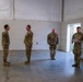 Four military personnel in OCP (Operational Camouflage Pattern) uniforms are gathered inside a large, sparse facility with white walls and a fabric/tent-style ceiling. One individual on the right appears to be reading from a document, while the others stand at ease facing him. The setting appears to be a temporary or semi-permanent military structure with a concrete floor.