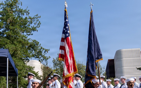 A U.S. Navy color guard ceremony is taking place outdoors on a clear, sunny day. Sailors in dress white uniforms are presenting the American flag and a Navy service flag, while rifle bearers stand at attention. A formation of additional Navy personnel stands in the background alongside a display of state flags.