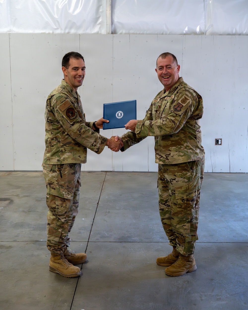 Two U.S. Air Force personnel in OCP (Operational Camouflage Pattern) uniforms are shaking hands while jointly holding a blue certificate or award folder bearing the U.S. Air Force seal. The ceremony appears to be taking place inside a hangar or large indoor facility with a concrete floor and white walls. Both individuals are smiling, suggesting a positive recognition or award presentation event.