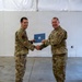 Two U.S. Air Force personnel in OCP (Operational Camouflage Pattern) uniforms are shaking hands while jointly holding a blue certificate or award folder bearing the U.S. Air Force seal. The ceremony appears to be taking place inside a hangar or large indoor facility with a concrete floor and white walls. Both individuals are smiling, suggesting a positive recognition or award presentation event.