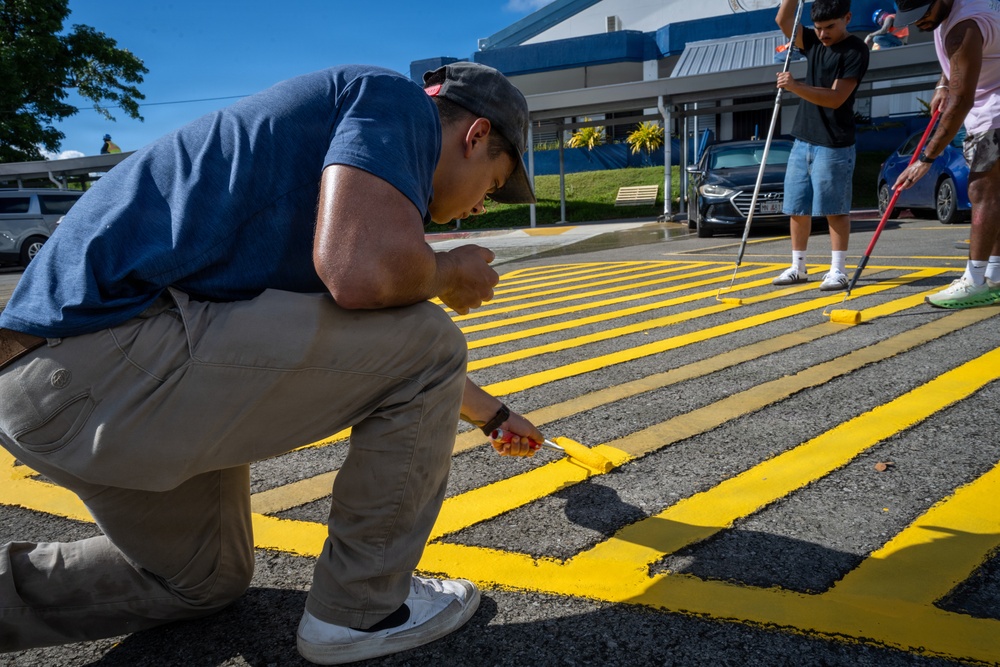 A group of people are painting bright yellow stripes on an asphalt surface, likely a parking lot or crosswalk area. The foreground shows a man crouching close to the ground applying paint with a small brush for detail work, while others in the background use long-handled rollers. The setting appears to be near a commercial or institutional building on a clear, sunny day.