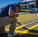 A group of people are painting bright yellow stripes on an asphalt surface, likely a parking lot or crosswalk area. The foreground shows a man crouching close to the ground applying paint with a small brush for detail work, while others in the background use long-handled rollers. The setting appears to be near a commercial or institutional building on a clear, sunny day.