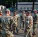 A group of six military personnel in OCP (Operational Camouflage Pattern) uniforms are gathered in a circle on a paved area at what appears to be a military installation or base. They appear to be engaged in an informal briefing or conversation outdoors. The setting includes military-style buildings, vehicles, and tall trees in the background.