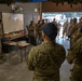 A military briefing is taking place inside what appears to be a garage or operations center, with a presenter pointing at a whiteboard covered in written notes. The group includes personnel wearing both U.S. military uniforms and Canadian Armed Forces uniforms, identifiable by Canadian flag patches on their sleeves. This appears to be a joint multinational military planning or coordination session.